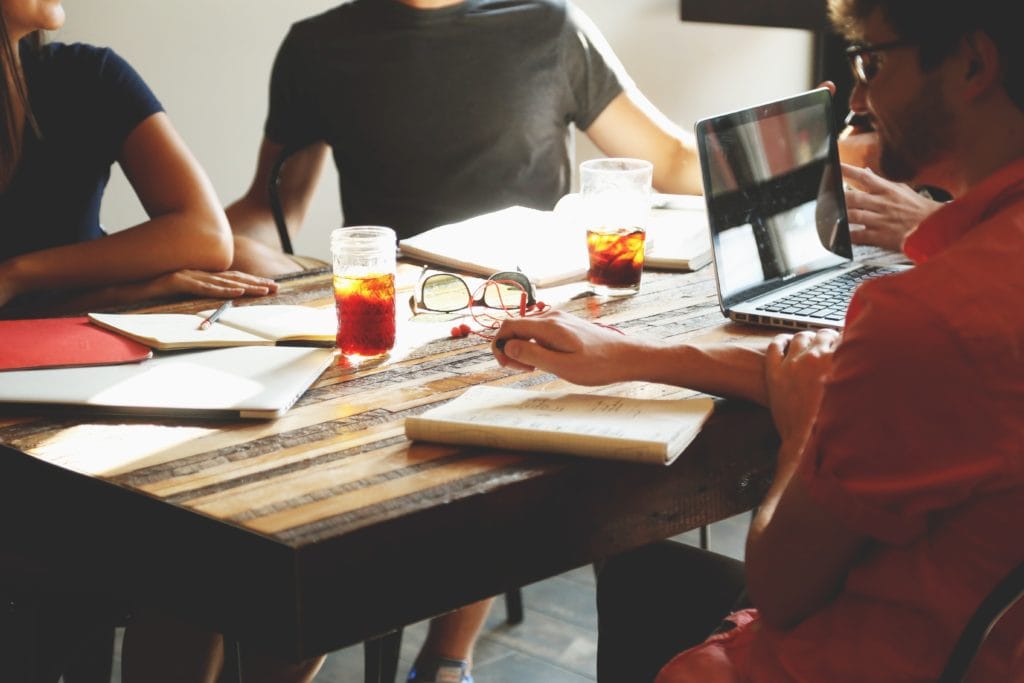 Group of people sitting around a table with notebooks and computers - used for a podcast about - How to Maximize Client Meetings When on a Business Trip