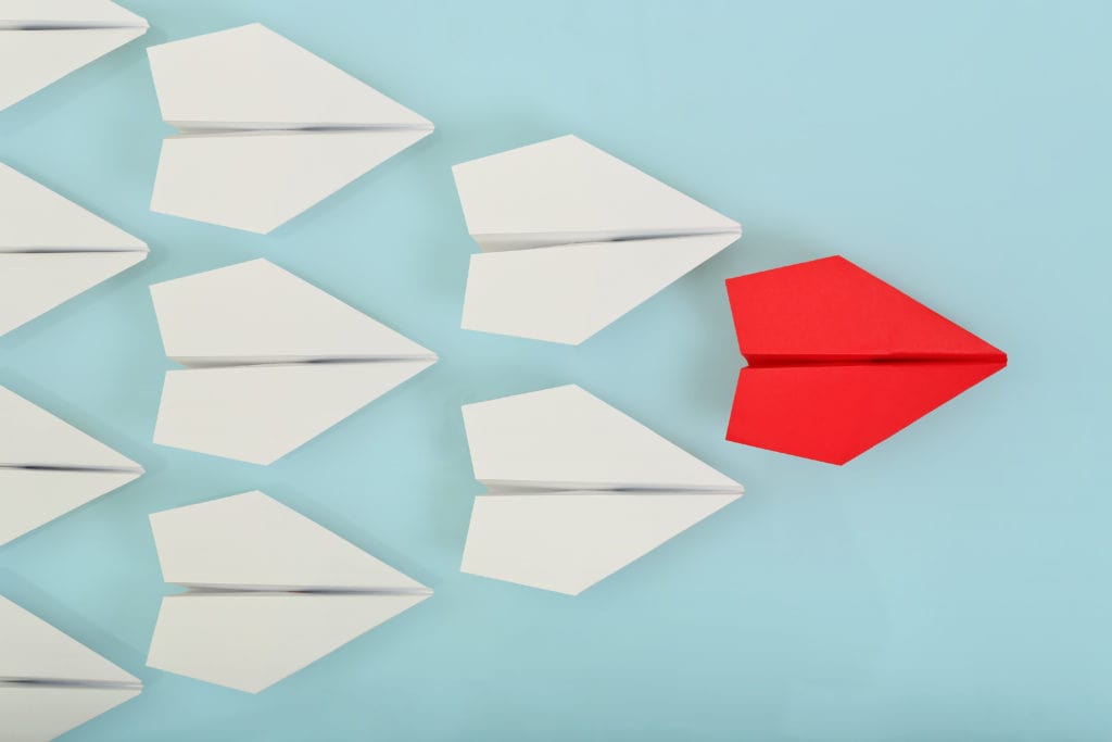 Red paper airplane leading white paper airplanes on a light blue background. Image is being used for a blog post about how the role of the leader has changed because of COVID-19.